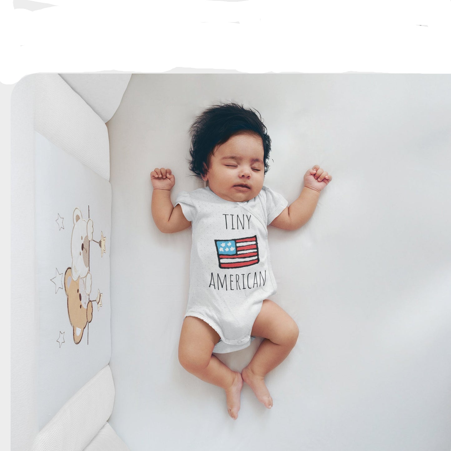 Baby wearing a onesie with 'Tiny American' text and an American flag, lying on a white surface.